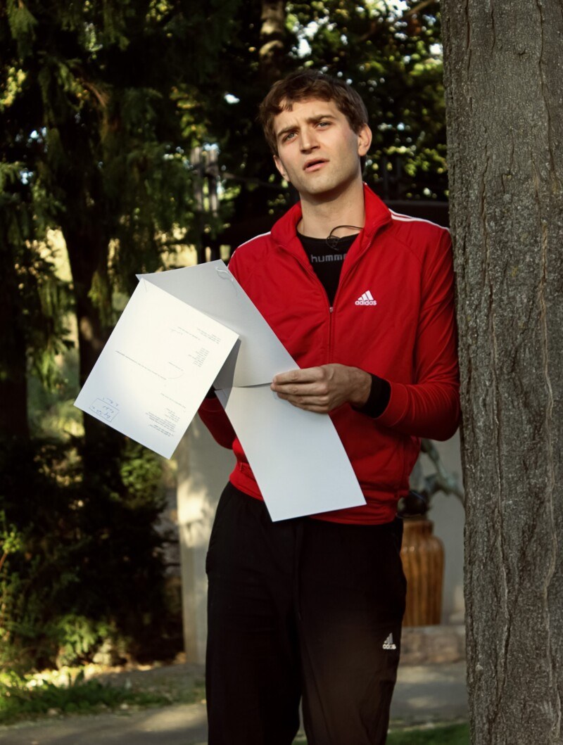 Hendrik Arns in roter Trainingsjacke neben einem Baum in einem Park, er liest im Nachmittagslicht laut von einem gefalteten weißen Blatt.