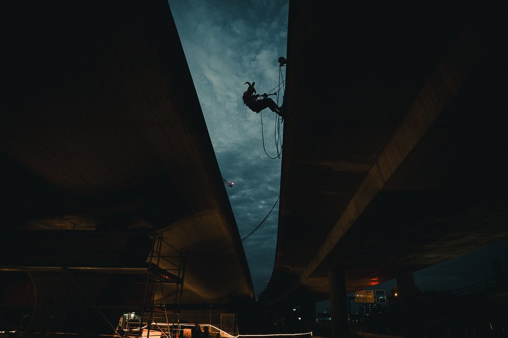 A figure abseils on a rope from the underside of a motorway bridge against a dark teal night sky, work lights below.