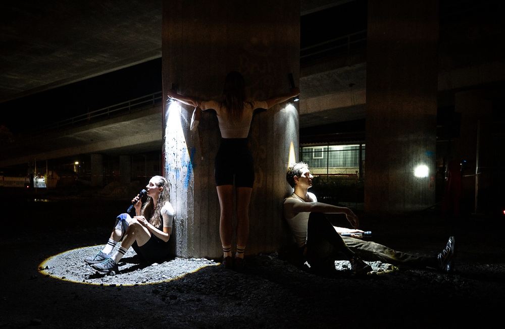Three performers under a concrete motorway bridge, one crouched in a spotlight, one standing, one seated on the ground.