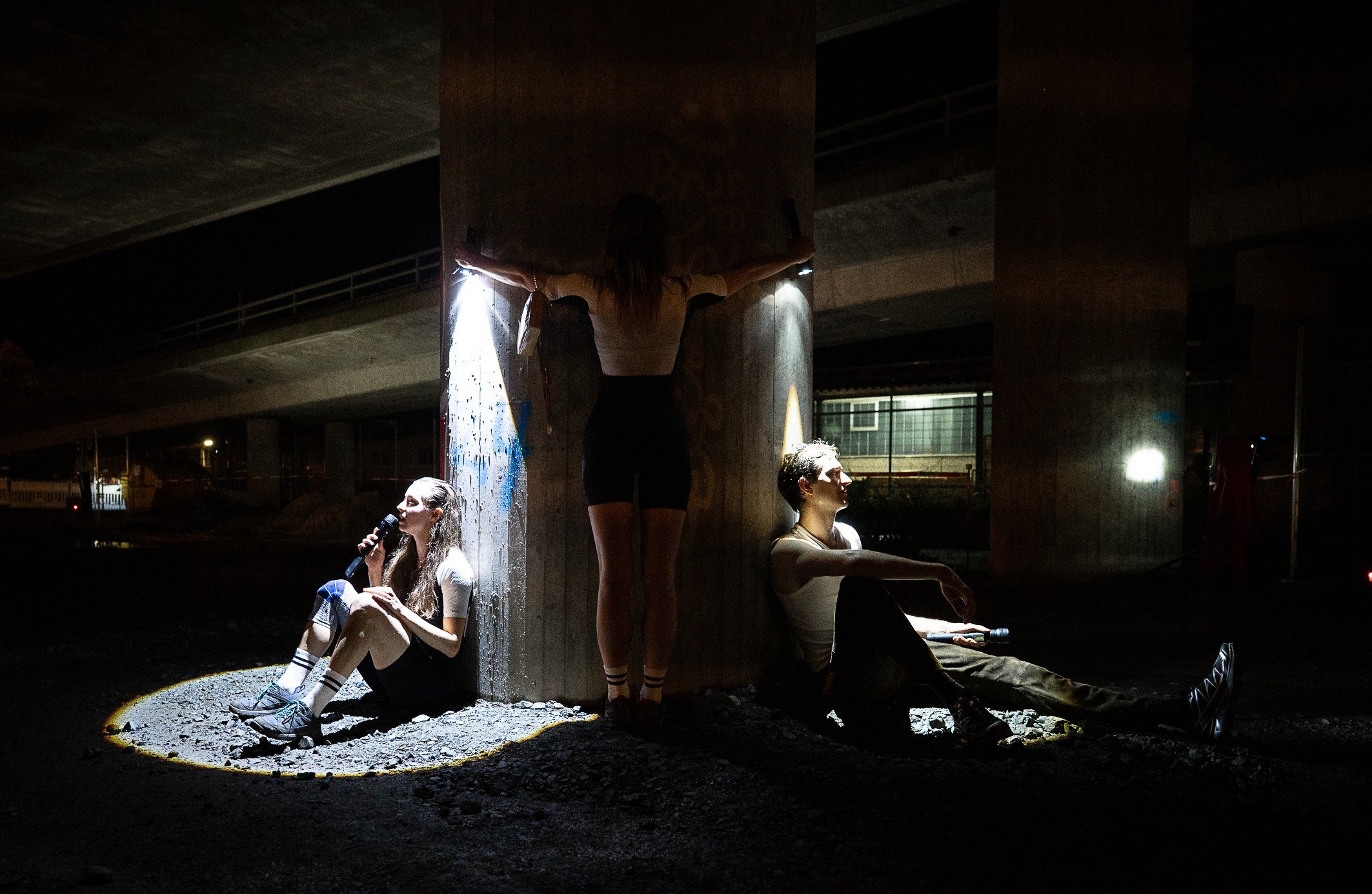 Three performers under a concrete motorway bridge, one crouched in a spotlight, one standing, one seated on the ground.