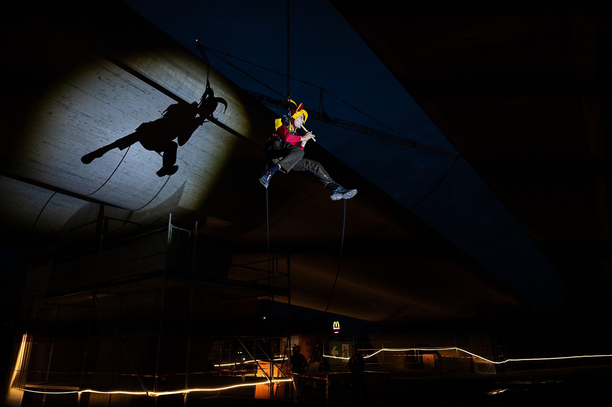 A performer in yellow and red hangs on ropes beneath the bridge deck at night.