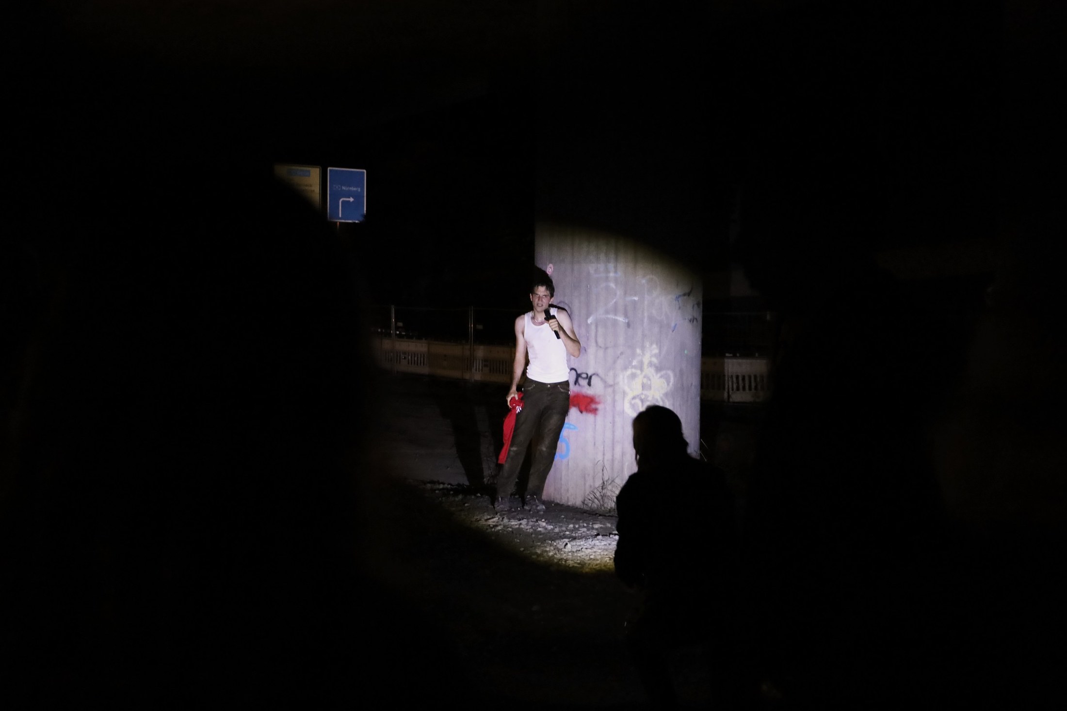 A performer stands alone in a dark underpass, lit from above, casting a long shadow.