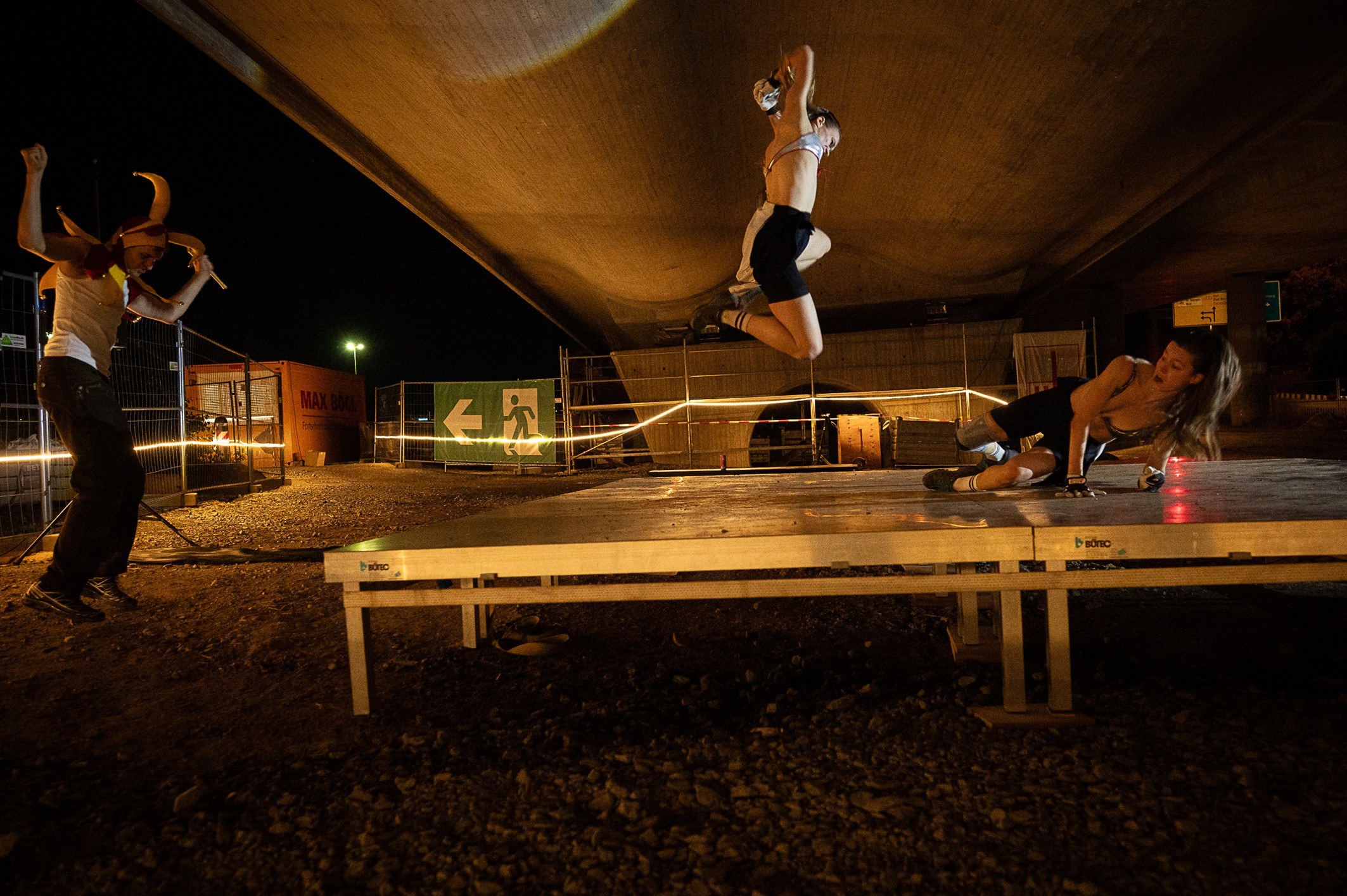 A performer leaps from a wooden platform under the bridge while others crouch on the ground.