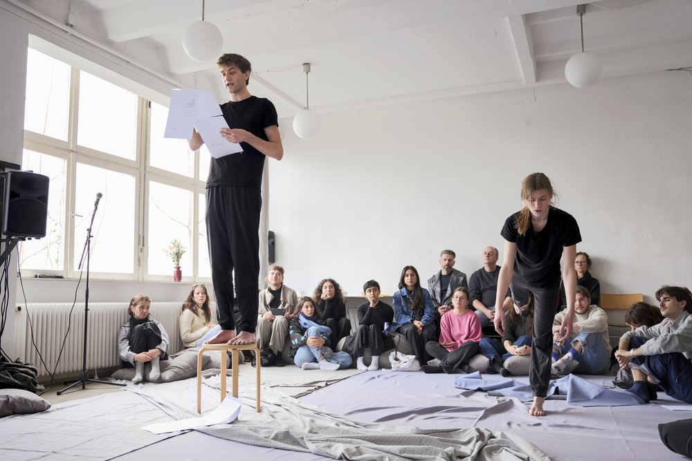 A performer stands reading from a sheet of paper in a bright room; the audience sits on the floor, a second performer at the right.
