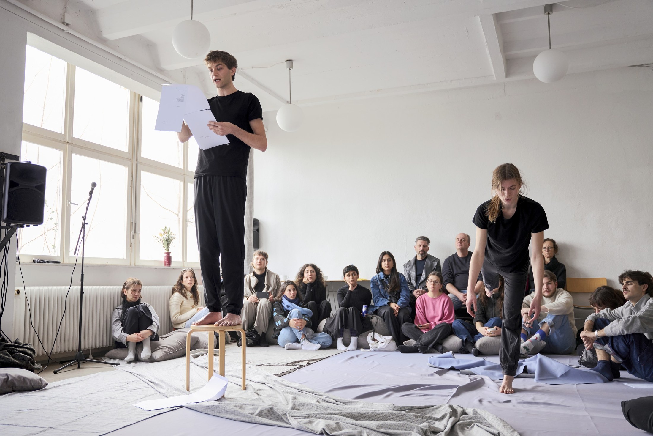 A performer stands reading from a sheet of paper in a bright room; the audience sits on the floor, a second performer at the right.