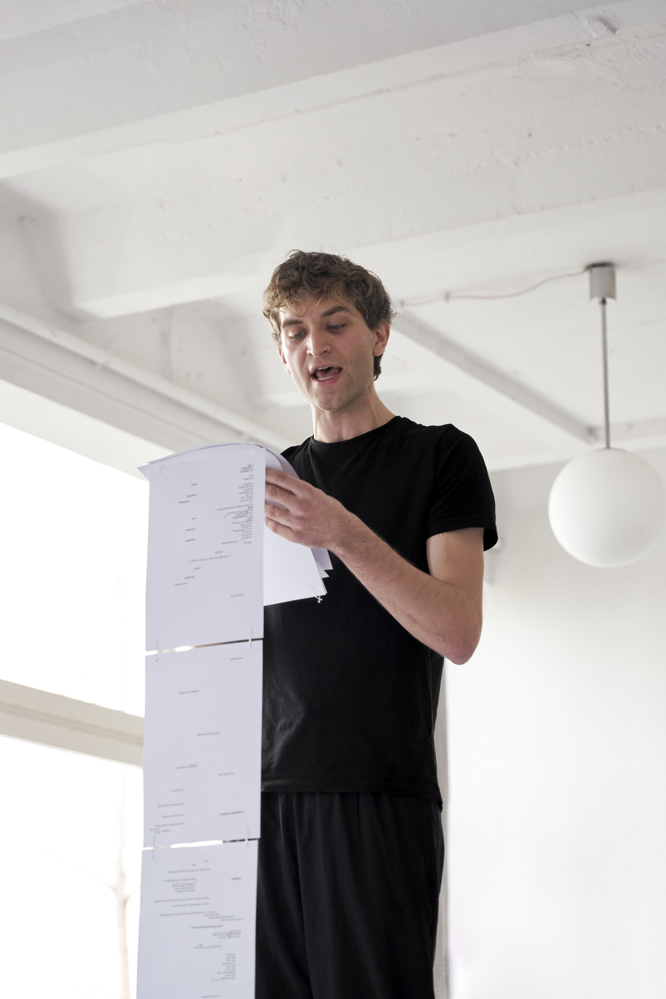 A performer in a black T-shirt reads aloud from a tall stack of white sheets in a bright room beneath a globe lamp.