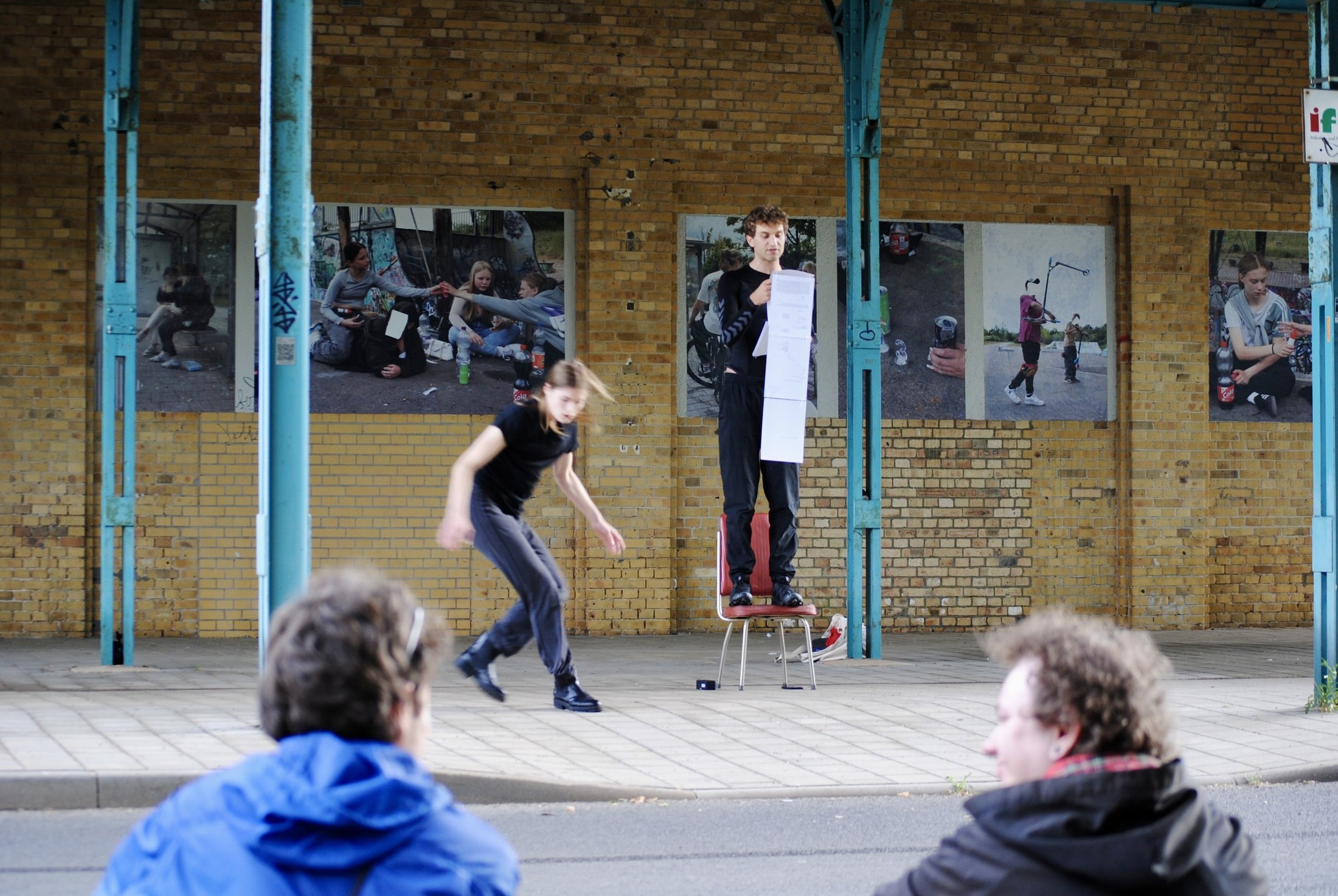Two performers on a low platform in front of a brick building hung with photographs; spectators in the foreground.