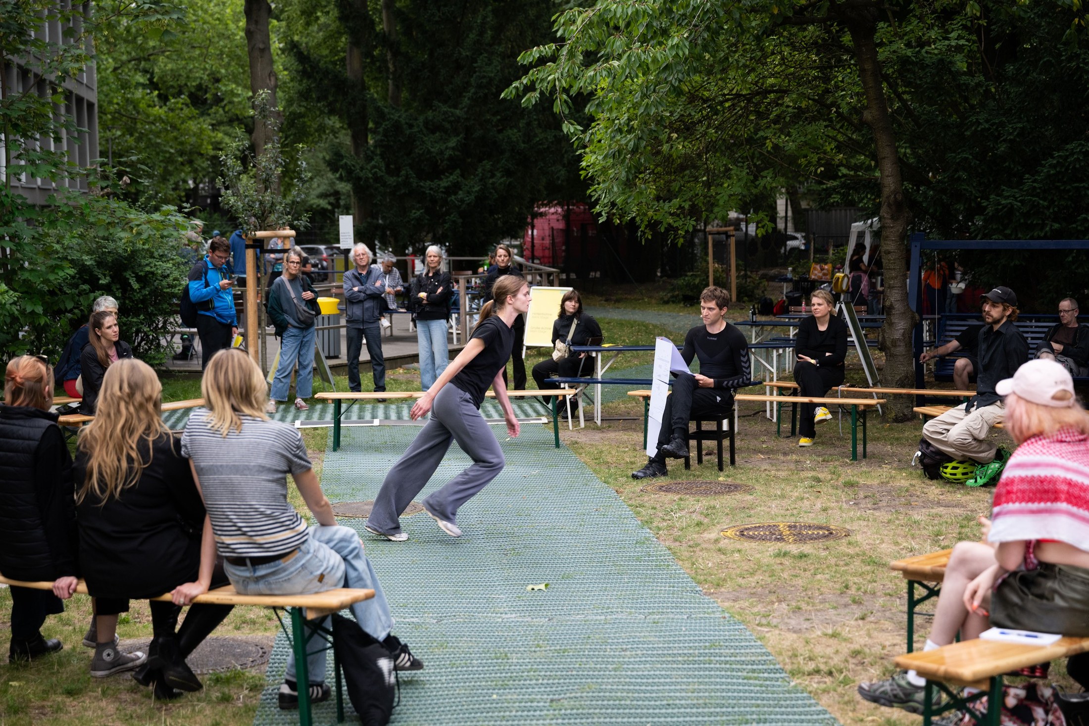 A performer runs across a lawn between two rows of benches in a park, the audience watching from both sides.