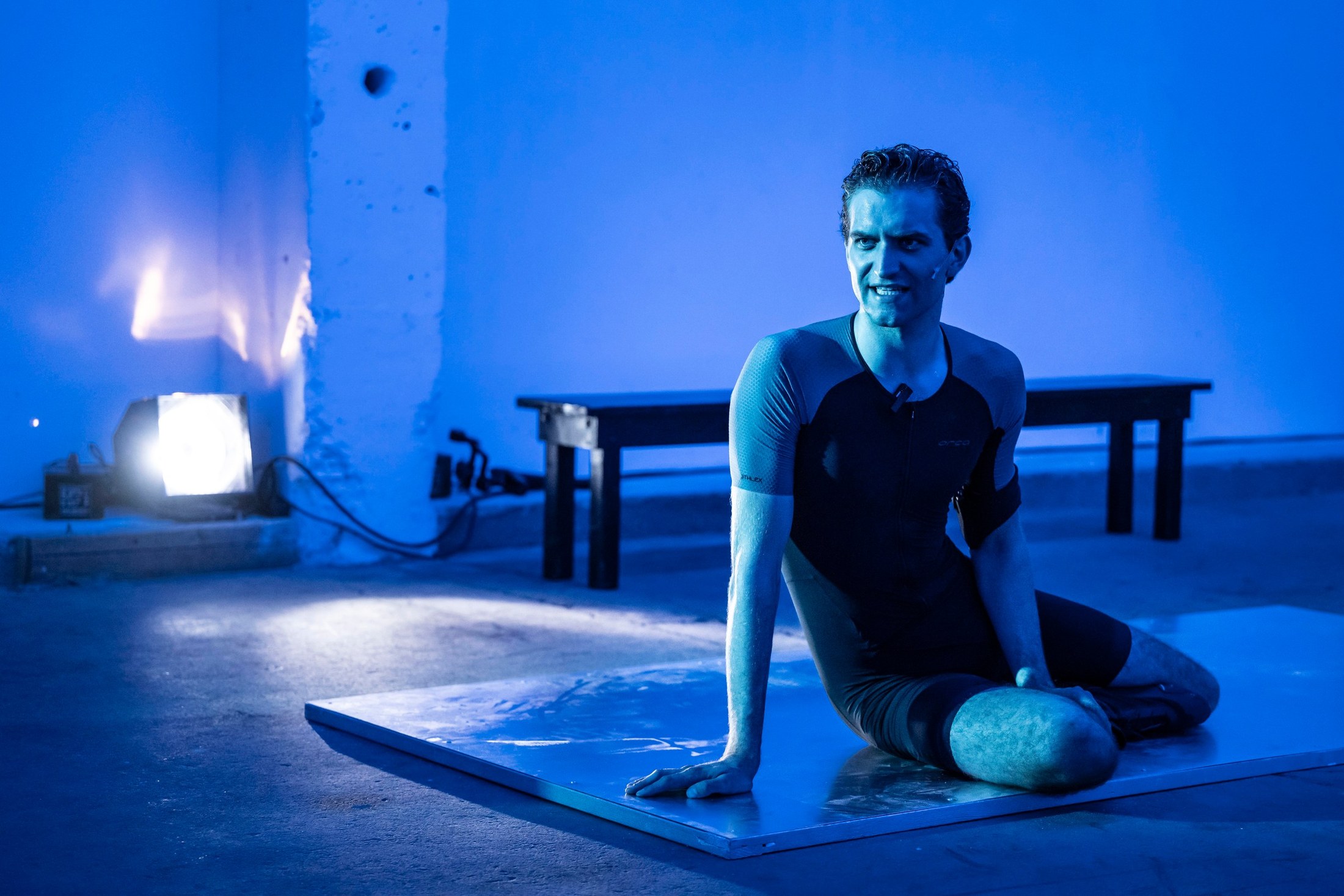 A performer kneels on the floor holding a pale egg-shaped object in blue light.