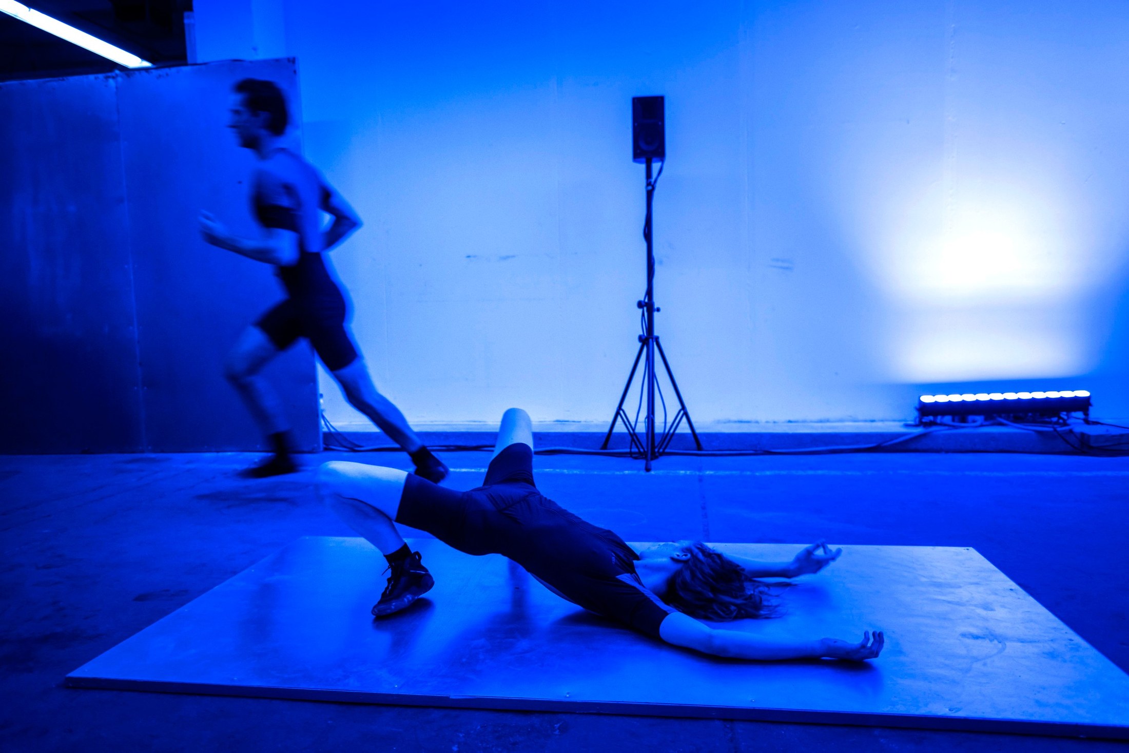 One dancer runs while another lies flat on the floor, blue light.