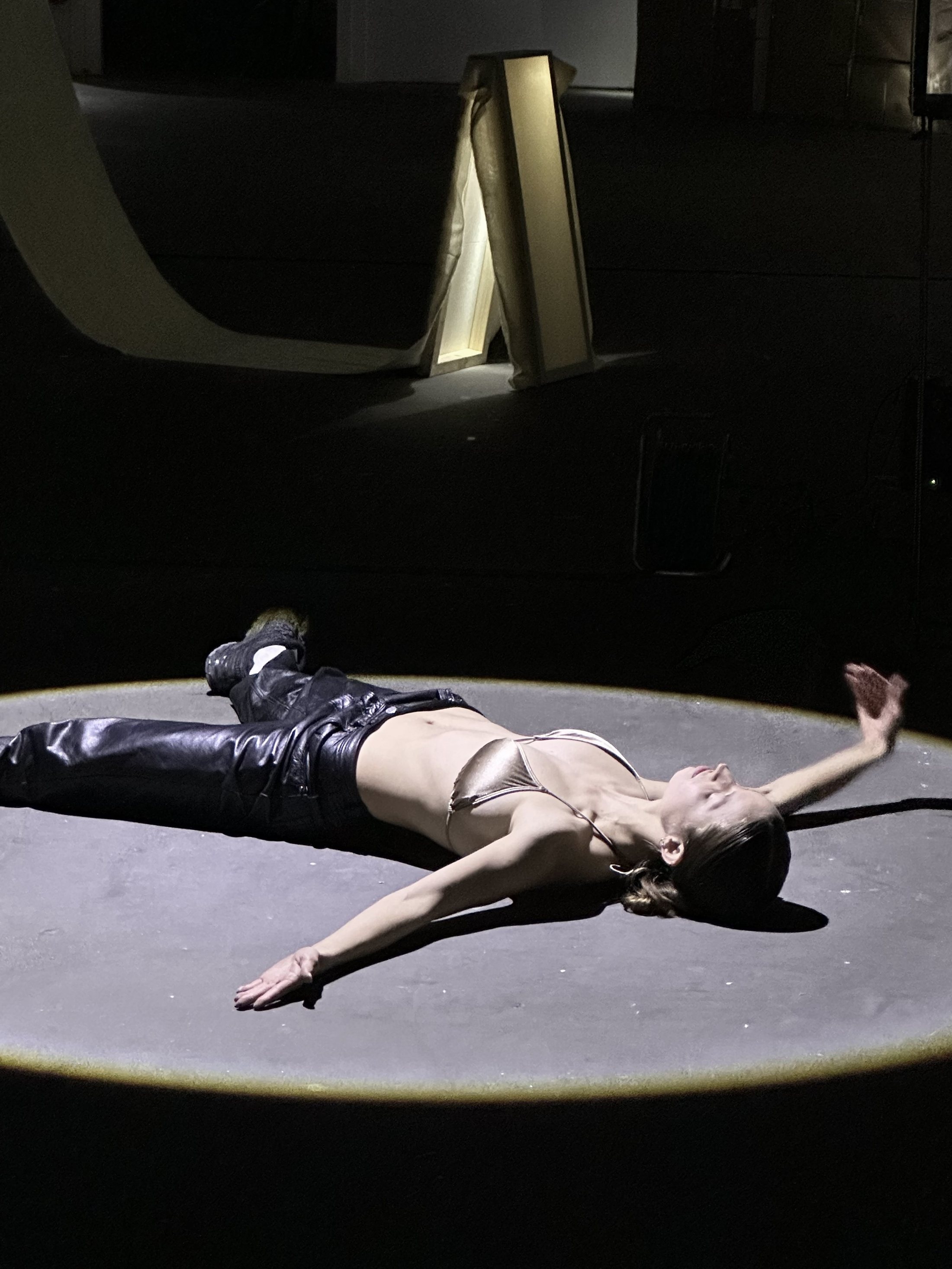 A performer in long black boots lies on their back in a spotlight on a dark floor, arms flung wide.