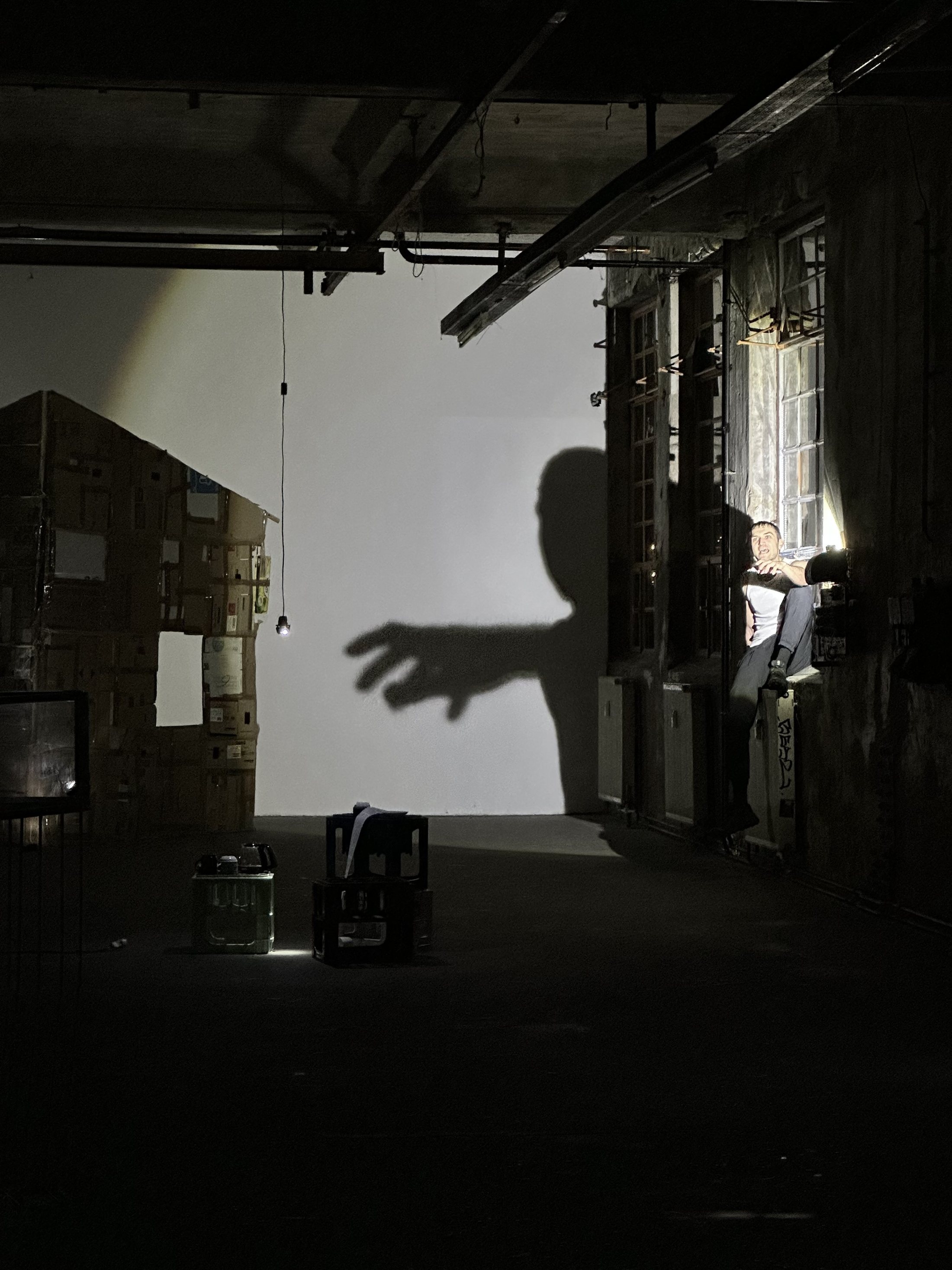 A huge shadow of an outstretched arm falls across the wall of a raw industrial room hung with sheets of paper.