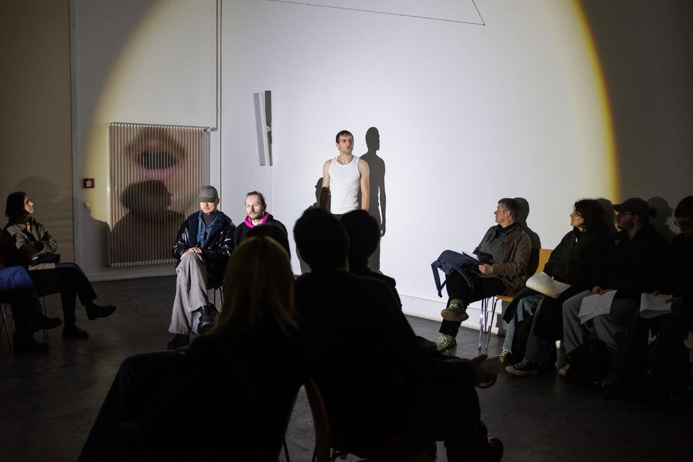 A performer stands against a white gallery wall while the audience sits on the floor around them.