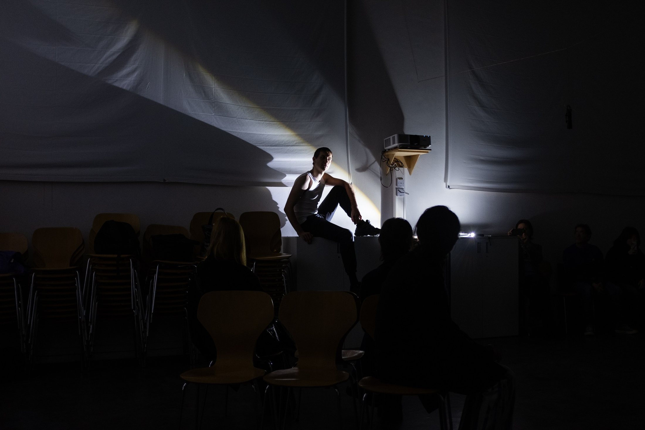 Silhouetted heads of the audience in the foreground; a performer sits in a lit doorway at the back of a dark hall.