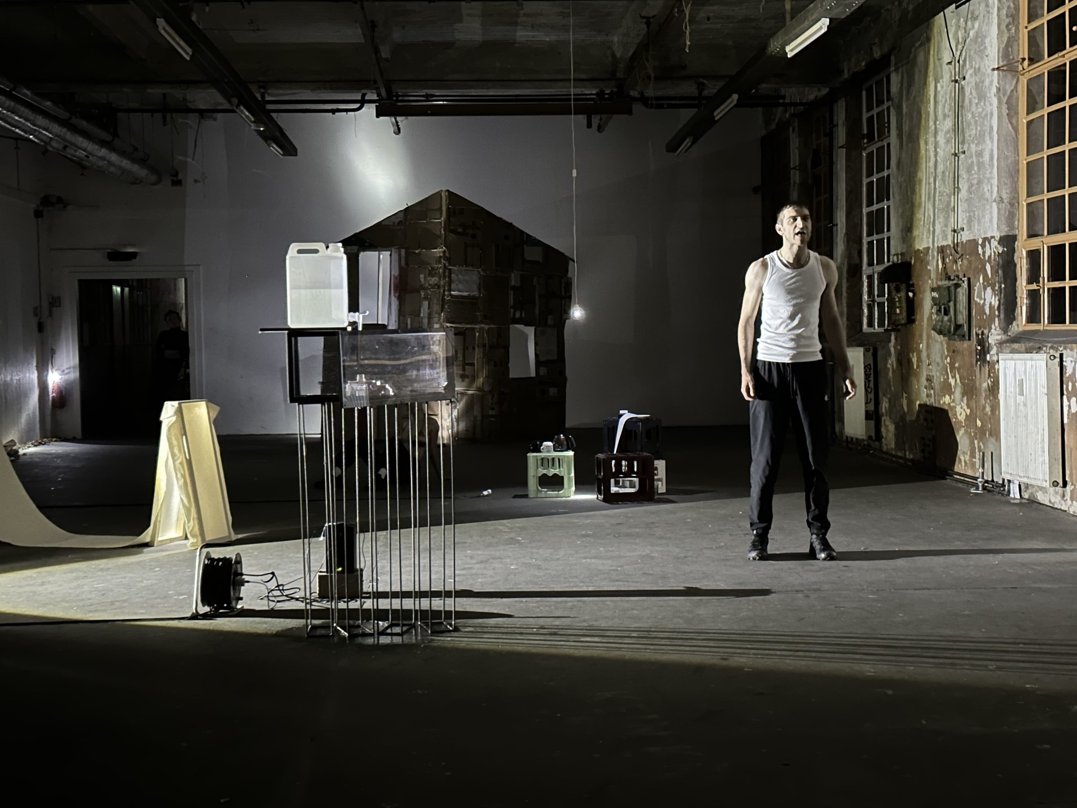 A performer walks through a raw industrial hall past a small hut and a hanging metal object.