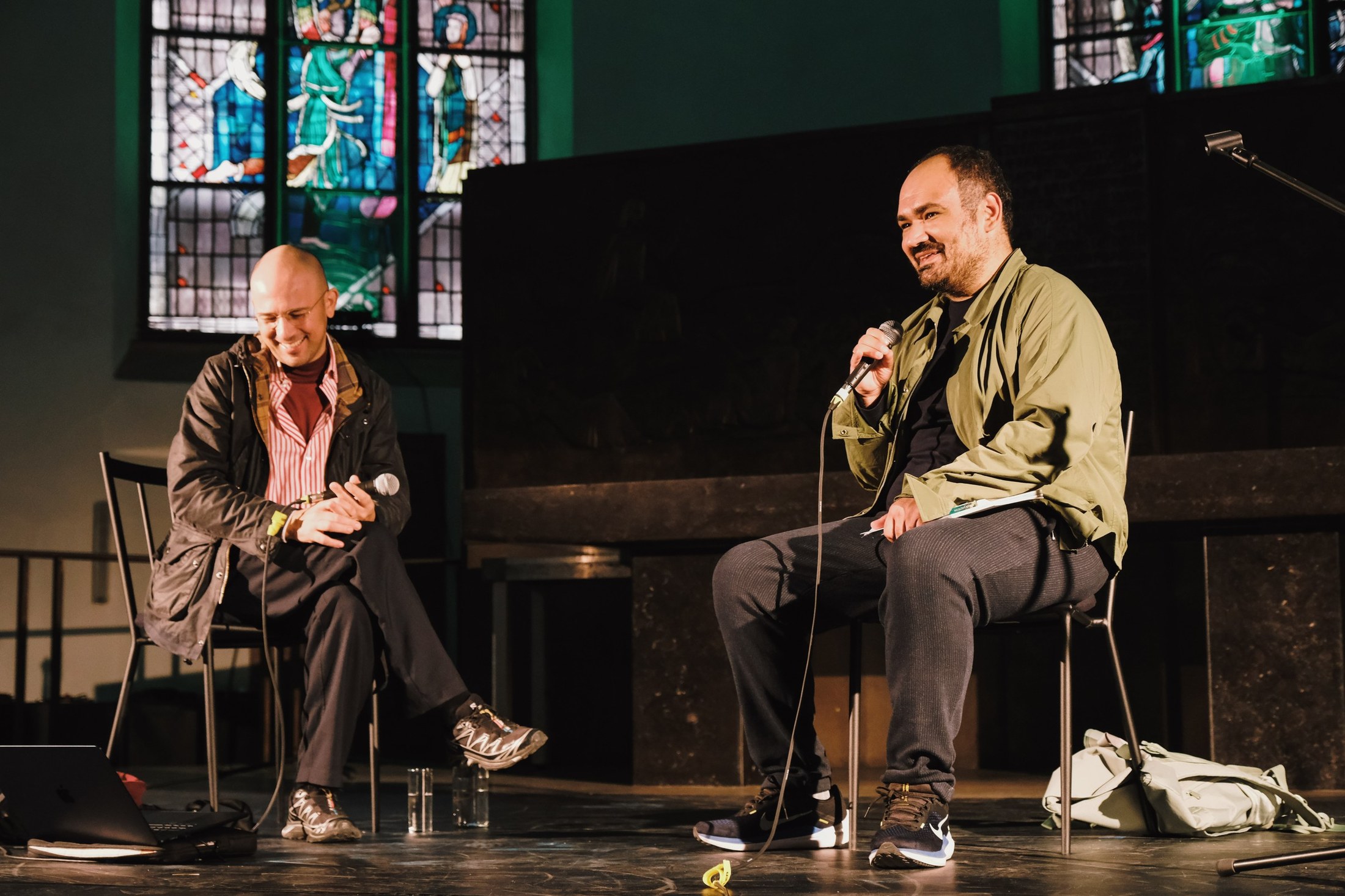 Two men sit talking with microphones on a stage in front of stained glass windows.
