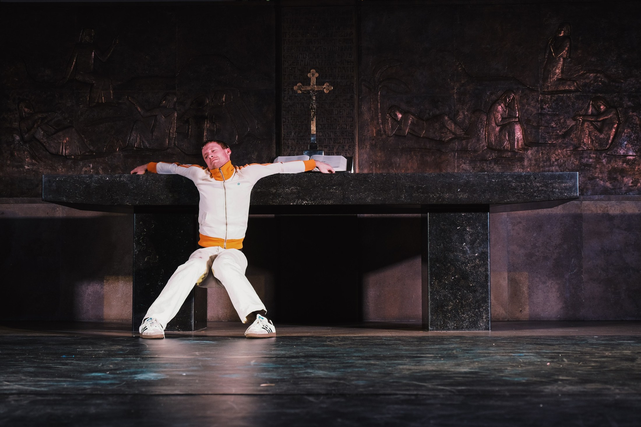 A performer in white stands with arms outstretched before a bronze altar relief and a cross.