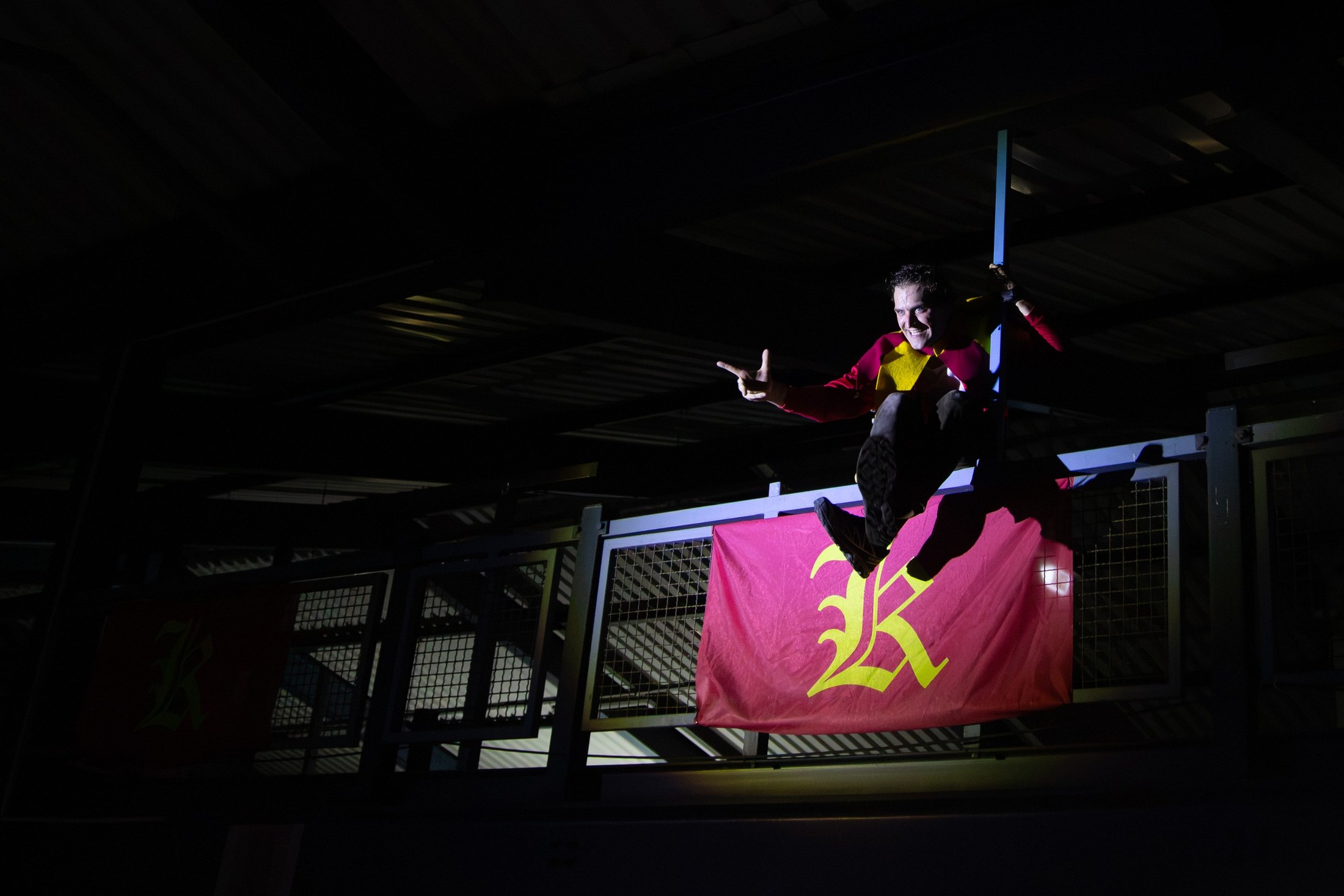 A performer in black and red jumps in a dark car park above a crimson banner bearing a yellow letter K.