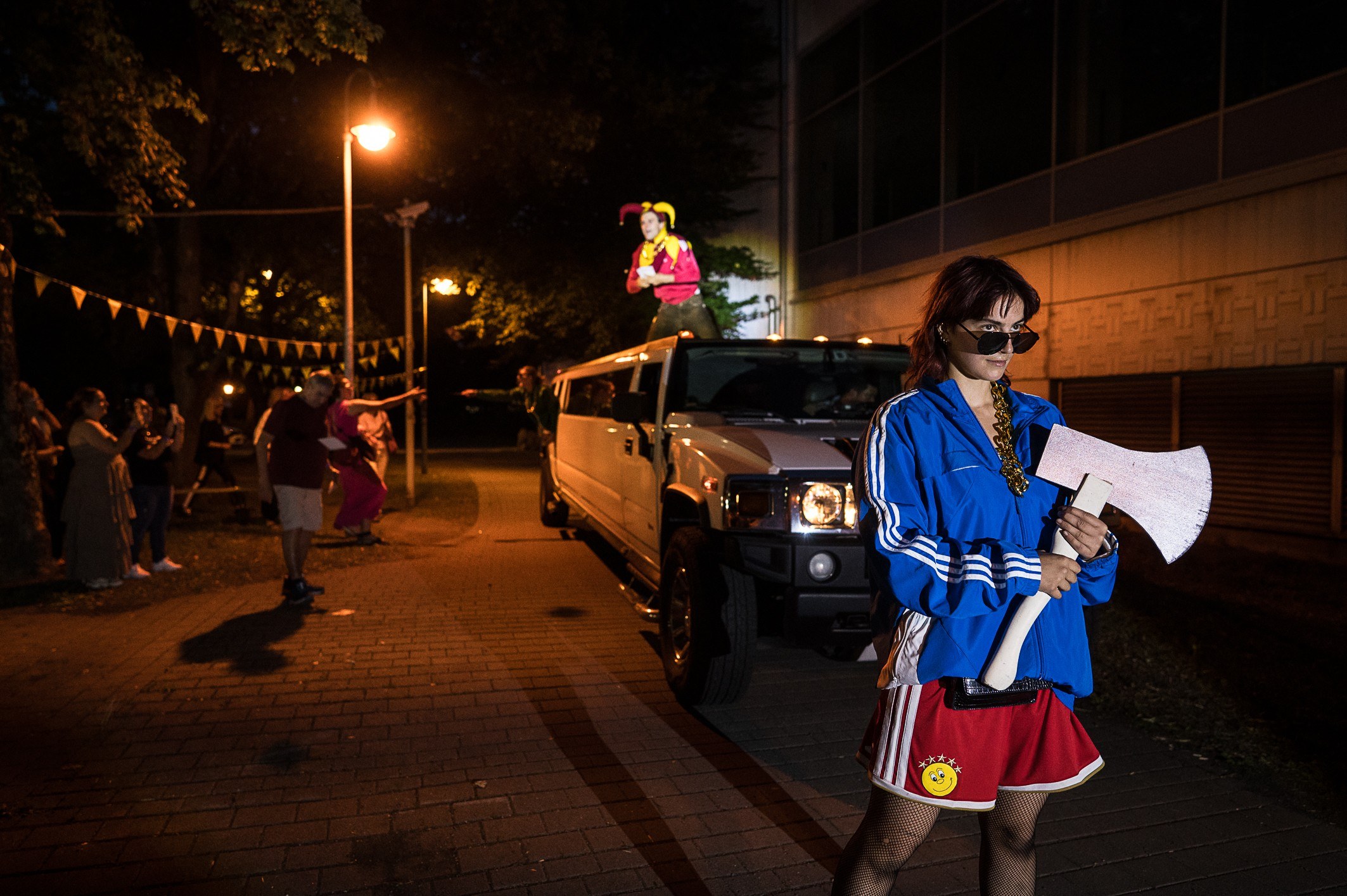 A performer in a blue tracksuit holds a megaphone beside a white stretch limousine at night, fairy lights strung overhead.