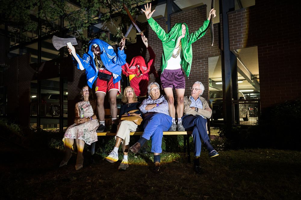 Three performers in bright tracksuits leap behind a park bench where five older audience members sit; one performer holds a cardboard axe. Night, brick building.