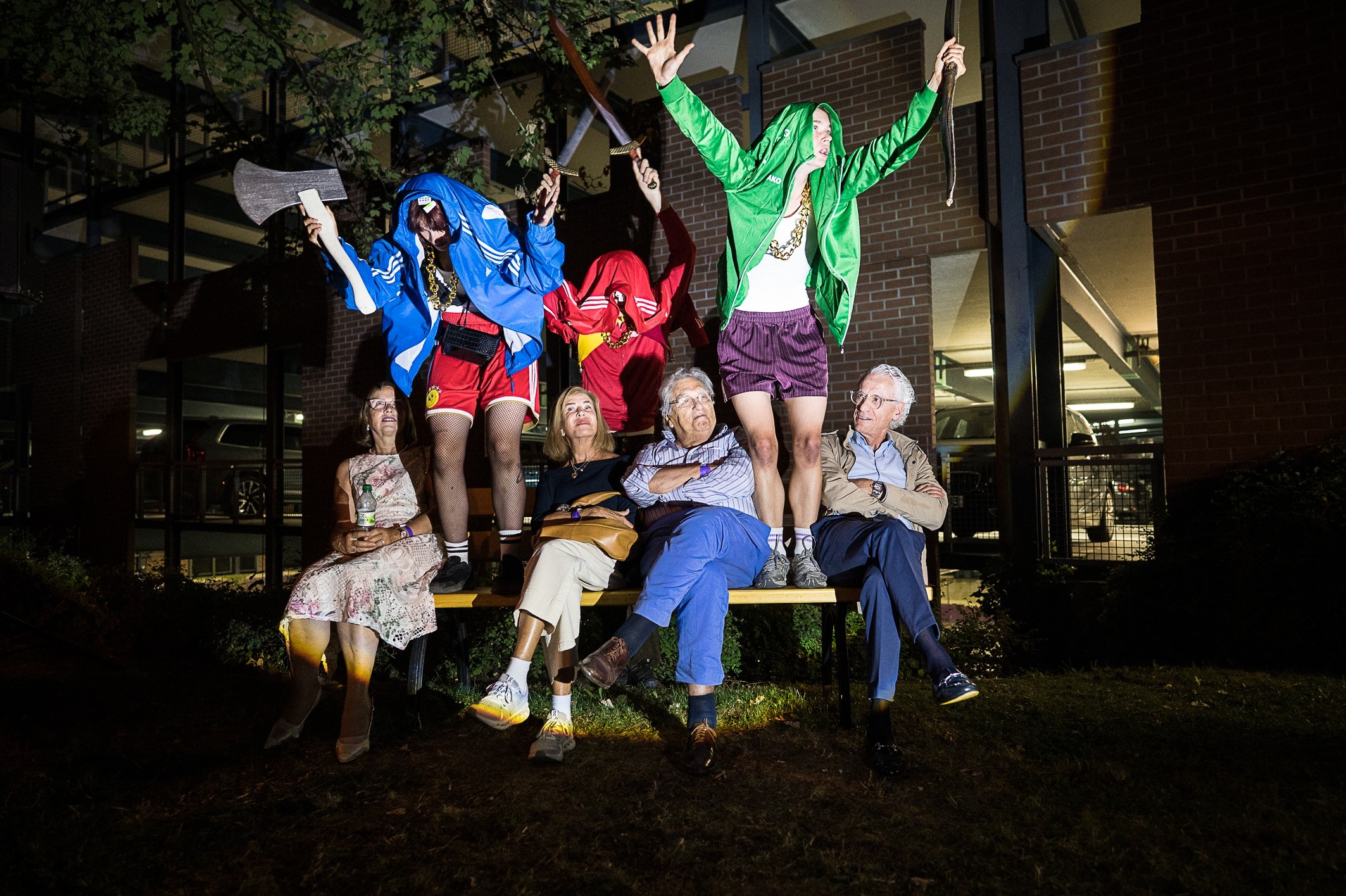 Three performers in bright tracksuits leap behind a park bench where five older audience members sit; one performer holds a cardboard axe. Night, brick building.