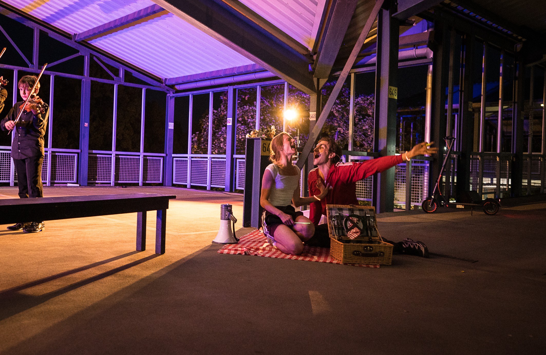 Performers under a violet-lit canopy roof; one stands on a long table, others sit on the floor beside a picnic blanket.