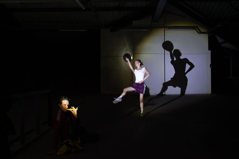 A performer with a ball throws a huge shadow onto the wall of a dark hall; a second figure kneels in the dark.