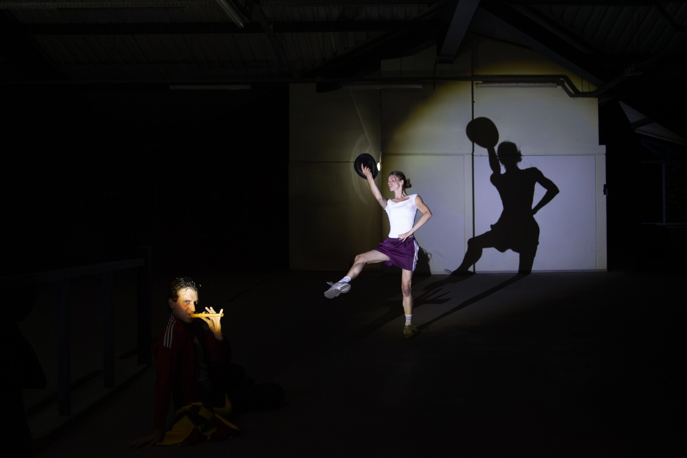 A performer with a ball throws a huge shadow onto the wall of a dark hall; a second figure kneels in the dark.